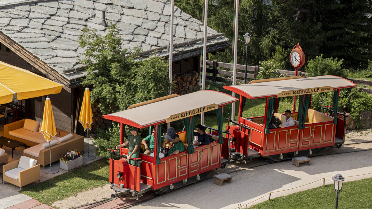 Le Riffelalp Tram, le plus haut tramway d'Europe, passant devant les Matterhorn Rooms du Riffelalp Resort 2222m à Zermatt, avec le Cervin et les Alpes suisses en toile de fond, symbole d'un séjour de luxe en montagne.